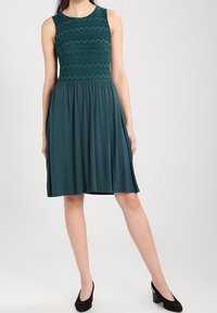 Robe sans manches en teal avec un motif zigzag texturé sur le buste, jupe fluide et tissu lisse. Associée à des talons noirs.