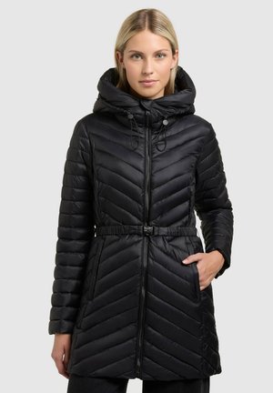 Kvinde iført en sort quiltet hættejakke med en lynlås foran, bælte i taljen og den ene hånd i sidelommen, stående mod en ensfarvet baggrund.