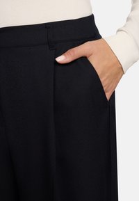 Pantalon noir à finition texturée, doté d'une poche latérale et d'une taille ajustée. Une main repose dans la poche, mettant en valeur le design.