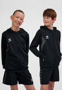 Schwarze Sportjacken mit Kapuzen, grauen Akzenten und Reißverschluss, kombiniert mit schwarzen Shorts mit Chevron-Muster.