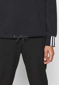 Top noir à manches longues avec un cordon de serrage, doté de quincaillerie dorée et d'une taille texturée. Les poignets ont trois rayures blanches.
