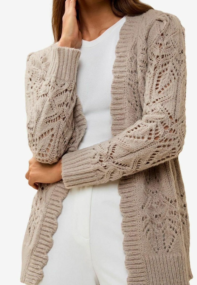 Femme portant un cardigan beige en tricot avec des motifs en dentelle par-dessus un top blanc et un pantalon blanc, main touchant son visage et bras croisés.