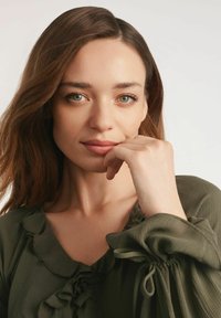 Oliwkowa bluzka z falbaną przy dekolcie i bufiastymi rękawami, wykonana z lekkiego materiału. Modelka delikatnie opiera dłoń na brodzie, patrząc w przód.