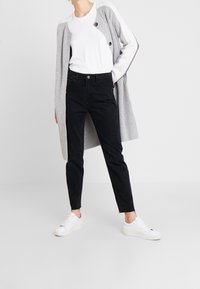 Jeans negros de talle alto con pierna ajustada, combinados con una camiseta blanca y un cárdigan gris claro con mangas en contraste y botones grandes.