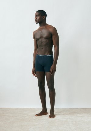 Homme musclé à la peau foncée, debout pieds nus, portant un boxer Björn Borg bleu marine, regardant vers la gauche sur un fond gris clair uni.