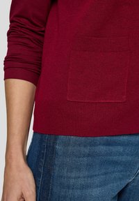 Roter Pullover mit langen Ärmeln und einer vorderen Tasche. Gerippte Struktur an Ärmelbündchen und Saum, kombiniert mit Jeans.