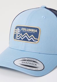 Gaiši zils snapback cepure, kas izgatavota no auduma ar režģa aizmuguri. Iespējo taisnstūrveida Columbia Sportswear logo tumši zeltītā krāsā ar ģeometrisku dizainu.
