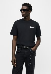 Άνδρας που φορεί μαύρα γυαλιά ηλίου, μαύρο μπλουζάκι "Dickies" και μαύρα τζιν με ασημένια σχέδια καρφιτσωμένα, στέκεται με το ένα χέρι στην τσέπη.