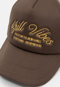 Brown baseball cap with gold embroidered text "Chill Vibes" and hashtags. Features a structured design and textured fabric with visible stitching.