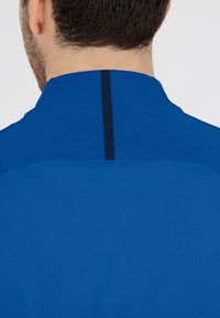 Maglietta sportiva blu con tessuto strutturato e finitura liscia. Una striscia scura verticale corre lungo il centro della parte posteriore del colletto.