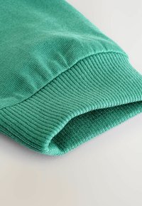 Närbild av en vik i ärmen på ett klart grönt plagg som visar en slät yta och ett ribbat mönster. Bakgrunden är neutral och mjuk.