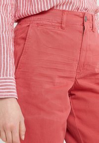 Osoba v korálově červených kalhotách a dlouhém rukávu se svislými pruhy v růžové a bílé barvě, s rukou odpočívající vedle kalhot.