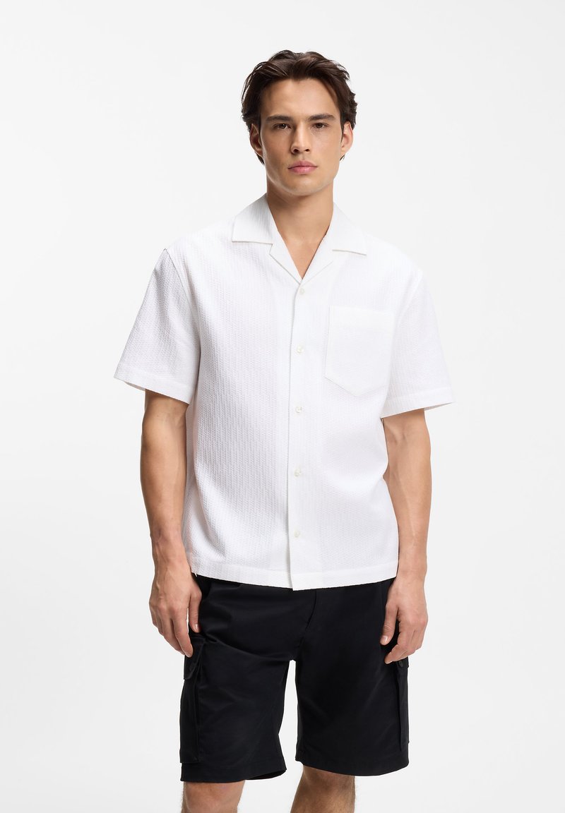 Νέος άνδρας που φοράει λευκό πουκάμισο με κοντά μανίκια και μαύρες cargo βερμούδες, μπροστά σε απλό λευκό φόντο.