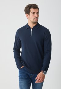 Camisola polo azul-marinho de mangas longas, com colarinho com fecho de correr, textura canelada e combinada com jeans azuis.