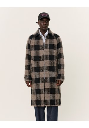 Long checkered coat in black and tan, made from textured fabric. Features a collar, button closure, and side pockets. Paired with dark jeans.