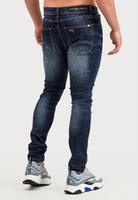 Tmavě modré džíny s úzkým střihem, zdobené skvrnami barvy, ošoupanými detaily a páskem s logem značky, doplněné šedými teniskami.