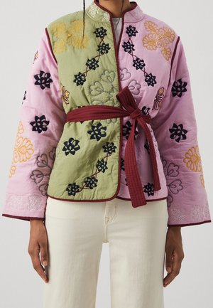 Chaqueta acolchada con paneles en rosa y verde, bordados florales, ribetes burdeos y un cinturón atado burdeos en la cintura, llevada con pantalones blancos.