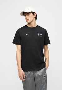 Νέος άνδρας που φοράει μαύρο t-shirt BMW Motorsport της Puma, ανοιχτό γκρι παντελόνια Puma και λευκό καπέλο Puma, στέκεται μπροστά σε μονόχρωμο φόντο.