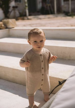 Maluch ubrany w beżowy, dziany kombinezon stoi na zewnątrz na kamiennych schodach w świetle dziennym.