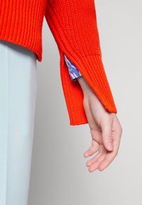 Pomarańczowy sweter z dzianiny o żebrowanej teksturze, z rozcięciem przy rękawie. Niebieska koszula w wzory jest częściowo widoczna pod swetrem.