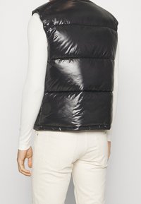 Schwarze ärmellose Steppweste aus glänzendem Material, mit gestepptem Design, Rückansicht, mit einem runden Stoffaufnäher an der Seite. Über einem hellen Oberteil getragen.