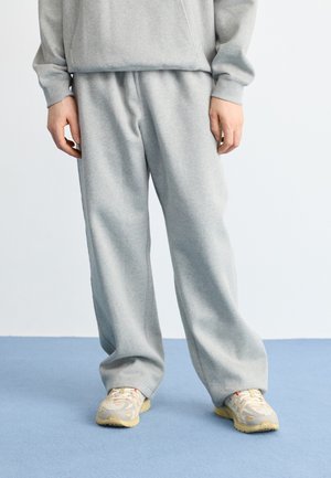 Pantalones de chándal gris claro, confeccionados con material suave, con un corte relajado y una cintura elástica. Combinados con zapatillas beige. Fondo azul.