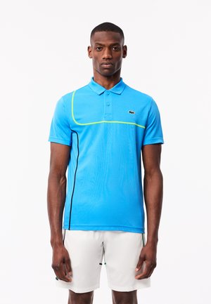 Niebieska koszulka polo o slim fit, z teksturowanej tkaniny, z żółtymi akcentami wzdłuż szwów oraz logo krokodyla na lewej piersi. W zestawie z białymi szortami.