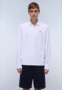 Polo blanca de manga larga con cuello y tres botones, con un pequeño logo en el pecho, combinada con pantalones cortos oscuros.
