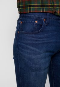 Jeans de mezclilla azul oscuro con un ajuste slim, diseño de cinco bolsillos y acentos de hardware en tono cobre. Textura suave con ligeros detalles de desvanecimiento.