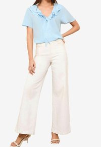 Blouse bleu clair avec col en V à volants et détail noué à l'avant. Associée à un pantalon blanc à jambes larges et des talons blancs.