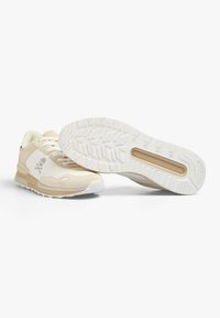 Baskets de sport beiges avec un mélange de matériaux lisses et texturés. Semelle extérieure en caoutchouc blanche avec un motif de bande de roulement, présentant un détail de logo en forme de crâne.