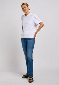 Camiseta blanca de manga corta con un bolsillo en el pecho, combinada con jeans ajustados azules y sandalias negras con detalle de hebilla.