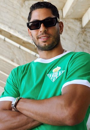 Groene T-shirt met korte mouwen en witte accenten, met een logo op de linkerborst. Zwarte vierkante zonnebril en een zilveren horloge worden gedragen.