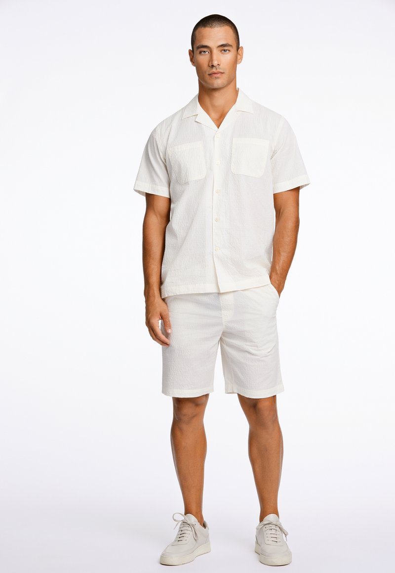 Heren twee-delig outfit in licht crèmekleurige stof; short-sleeve overhemden met twee voorzakken en bijpassende shorts, gecombineerd met witte sneakers.
