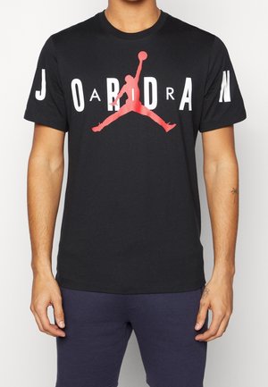 Mężczyzna ubrany w czarną koszulkę z czerwonym logo Jumpman i białym napisem "JORDAN", w zestawie z ciemnoniebieskimi szortami.