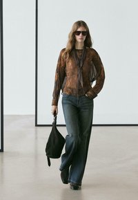 Brun mønstret langærmet bluse med bindebånd i halsen, kombineret med mørkeblå, vide jeans og en sort håndtaske, på en glat gulv.