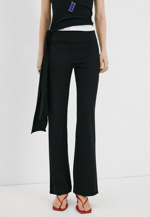 Žena oblečená v čiernych nohaviciach s rozšírenými nohavicami, čiernom tielku bez rukávov so zaväzovaním na boku a červených sandáloch na štvorcovom opätku, stojaca pred bielym pozadím.