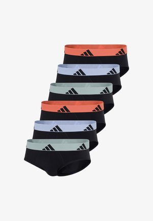 Athletischer dunkelhäutiger Mann steht barfuß da, trägt schwarze Slips mit einem orangen Adidas-Logo auf dem Bund, vor einem schlichten weißen Hintergrund.