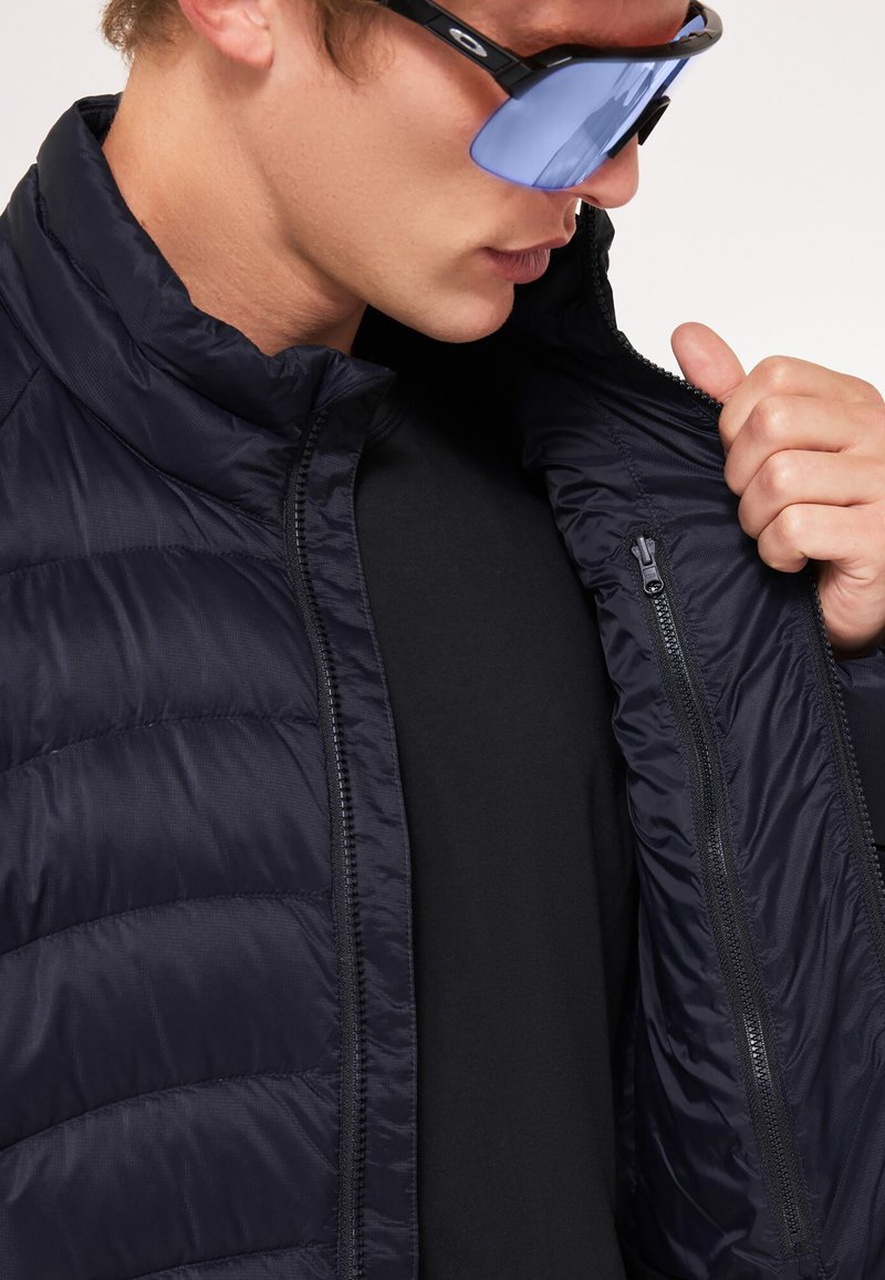 Chaqueta acolchada azul marino con patrón de acolchado diagonal, cremallera en la parte delantera y cuello alto. Llevada sobre una camiseta negra, con gafas de sol de tono azul.