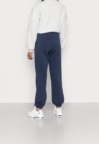 Marinblå joggers med elastiska muddars och midja, ihop med en ljusgrå sweatshirt, bärs med vita träningsskor på en enfärgad bakgrund.