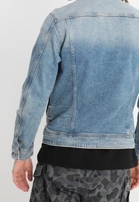 Person som bär en ljusblå denimjacka över en svart skjorta och grå kamouflagebyxor, sedd bakifrån.