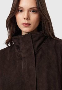 Giacca in camoscio marrone con collo alto e design minimalista; presenta cuciture eleganti e una texture morbida, concentrandosi su una silhouette pulita.