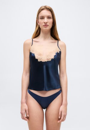 Nő tengerészkék szaténmelltartót visel bézs csipkés díszítéssel és hozzáillő tengerészkék bikini stílusú alsóneműt, áll egy egyszerű háttér előtt.