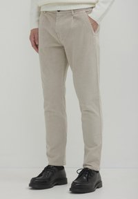 Pantaloni in corda beige indossati con scarpe nere con lacci e un maglione bianco a coste, con una mano in tasca su uno sfondo neutro.