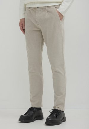 Pantaloni in corda beige indossati con scarpe nere con lacci e un maglione bianco a coste, con una mano in tasca su uno sfondo neutro.