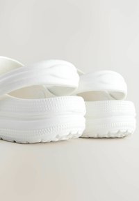 Weiße Slipper mit dicken, strukturierten Sohlen, glatten Riemen und einem abgerundeten Design. Leichtes Material und minimalistischer Stil sind deutlich erkennbar.