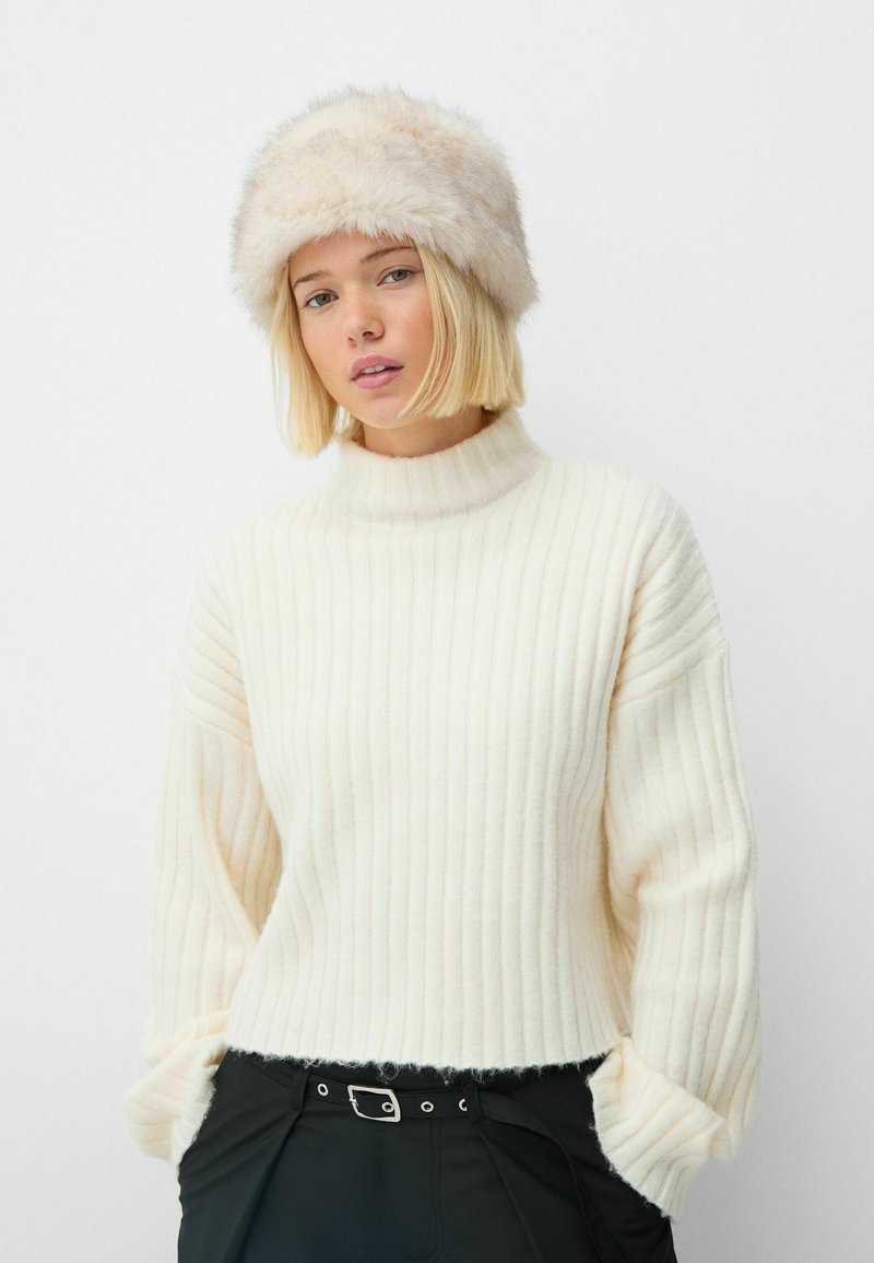Flauschiger Cremepullover mit gerippter Textur und hohem Kragen, kombiniert mit einem weichbeigen Fellhut, getragen mit schwarzen Hosen und einem Gürtel.