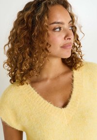 Suéter amarillo de punto con escote en V y mangas cortas. Textura suave y aterciopelada con borde acanalado en el escote y las mangas.