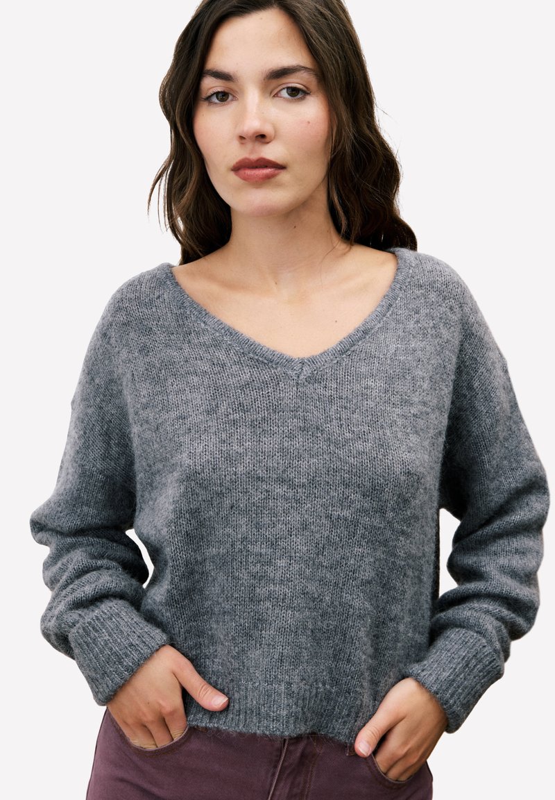 Vrouw met lang bruin haar, gekleed in een grijze gebreide V-halstrui en paarse broek, handen in de zakken, neutrale uitdrukking, witte achtergrond.