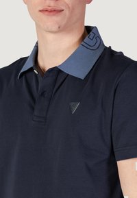 Polo shirt blu navy con colletto azzurro chiaro caratterizzato da un motivo geometrico. Include un piccolo logo a triangolo sul petto e una trama in morbido cotone.